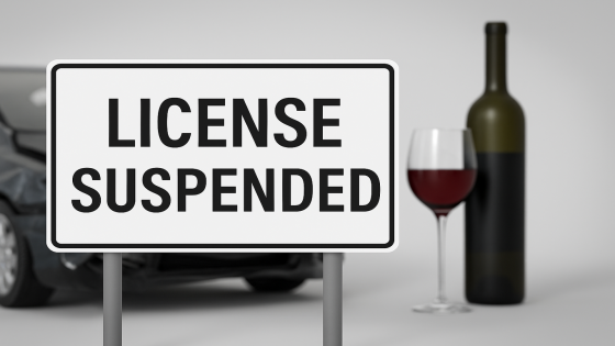 لافتة مكتوب عليها "LICENSE SUSPENDED" أمام سيارة محطمة وزجاجة نبيذ، ترمز إلى تعليق رخصة السياقة بسبب القيادة تحت تأثير الكحول